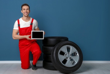 Photo of Young mechanic in uniform with tablet PC and car tires near color wall