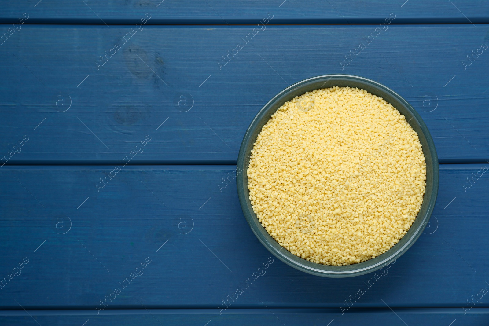 Photo of Bowl of raw couscous on blue wooden table, top view. Space for text