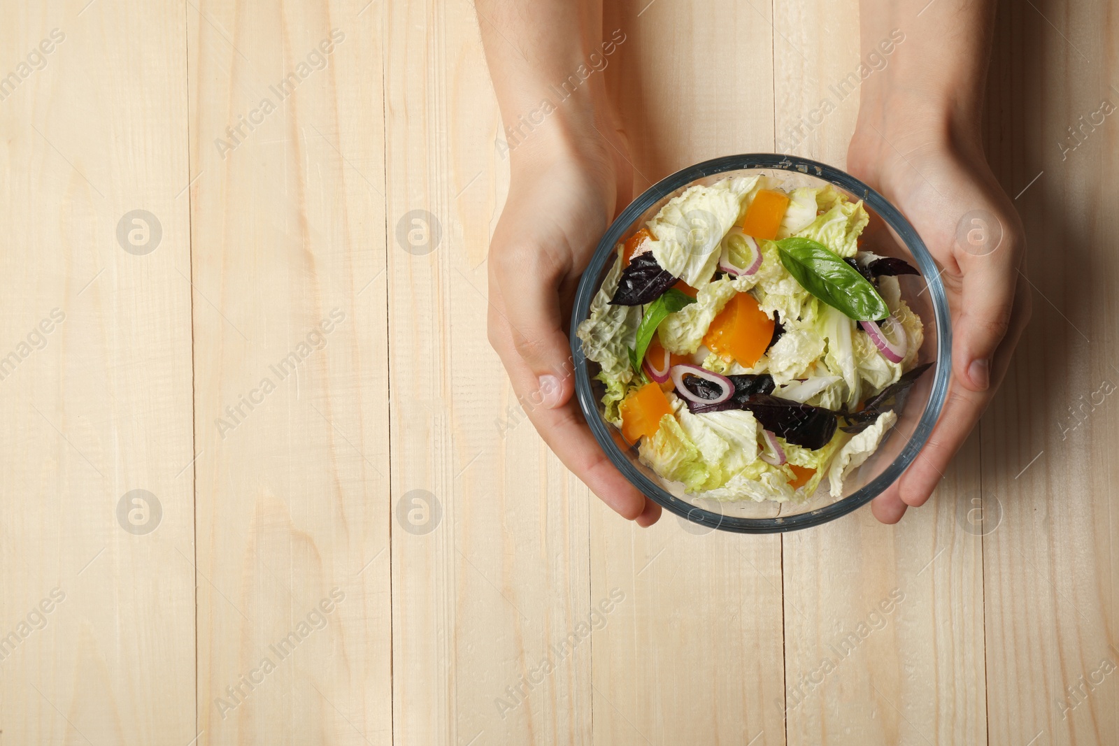 Photo of Woman holding bowl of delicious salad with Chinese cabbage, tomato and basil at wooden table, top view. Space for text