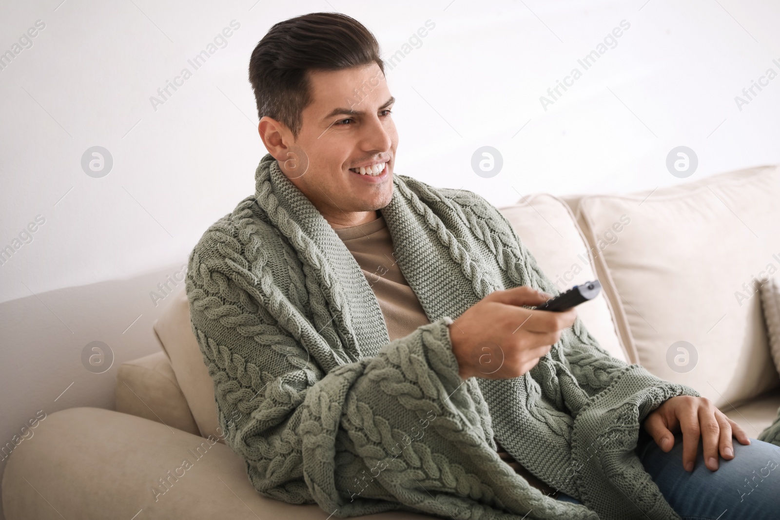 Photo of Man covered with warm green plaid watching television on sofa at home
