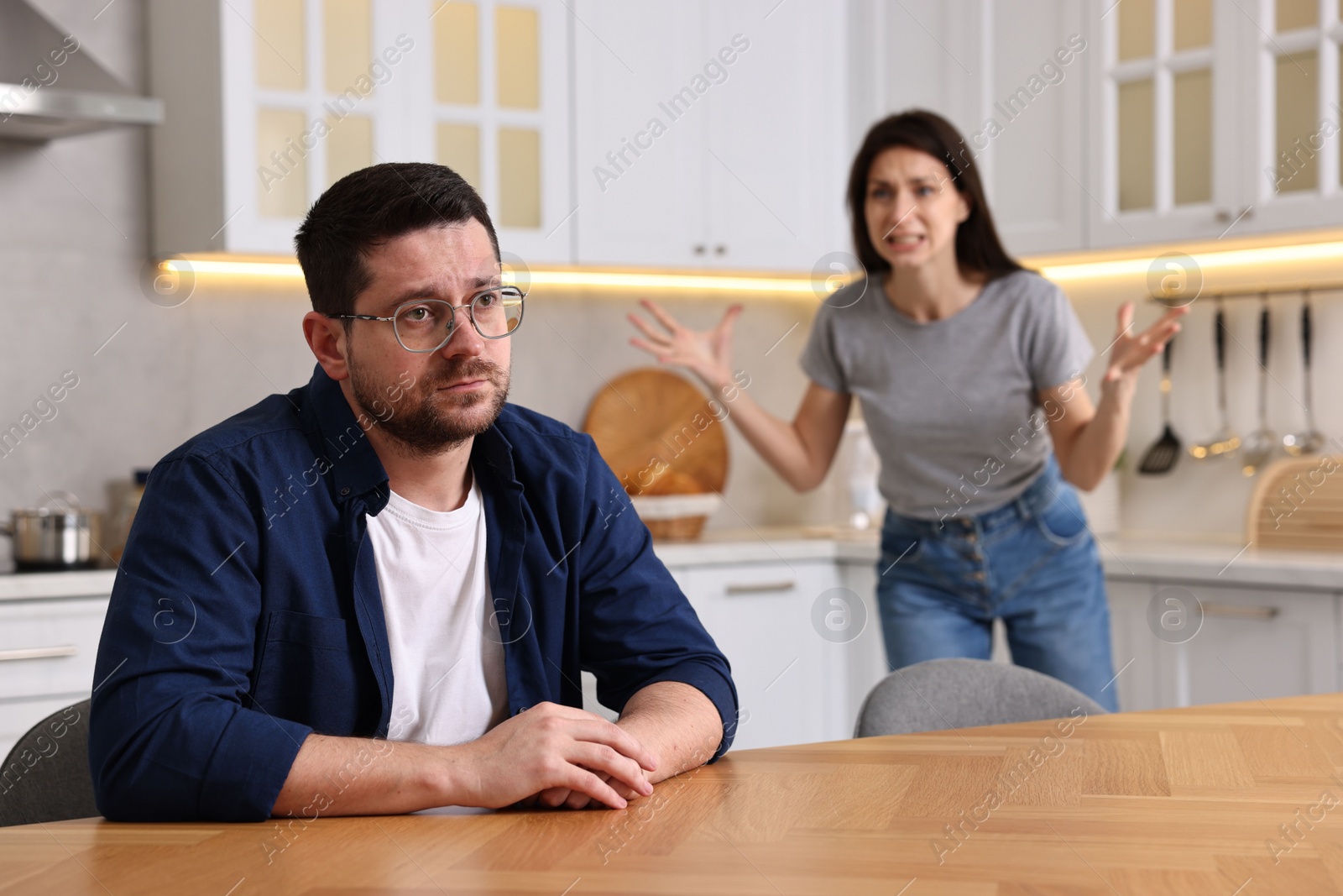 Photo of Sad husband sitting at table while his wife screaming at him in kitchen, selective focus. Relationship problems