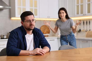 Sad husband sitting at table while his wife screaming at him in kitchen, selective focus. Relationship problems