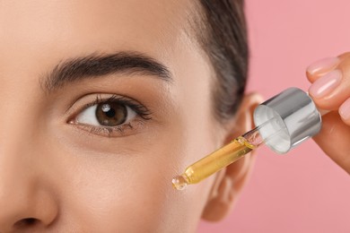 Young woman applying serum onto her face on pink background, closeup