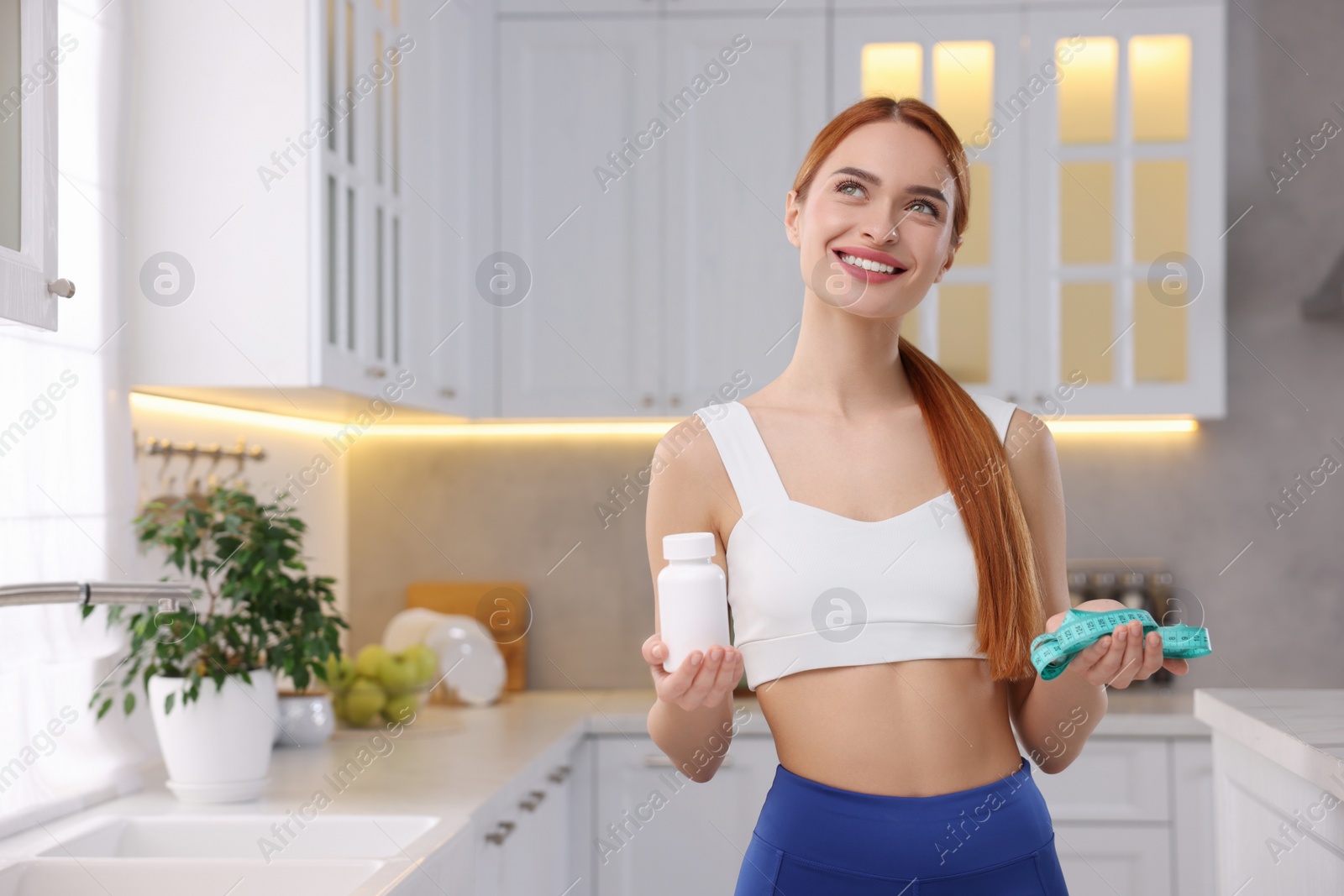 Photo of Happy young woman with bottle of pills and measuring tape in kitchen, space for text. Weight loss