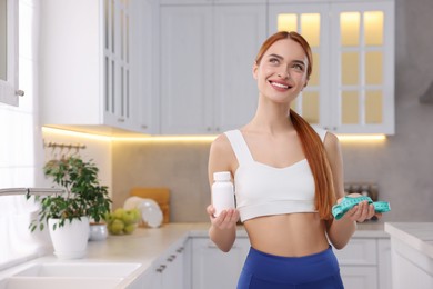 Happy young woman with bottle of pills and measuring tape in kitchen, space for text. Weight loss