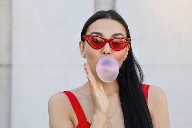 Beautiful woman in stylish sunglasses blowing gum near wall outdoors