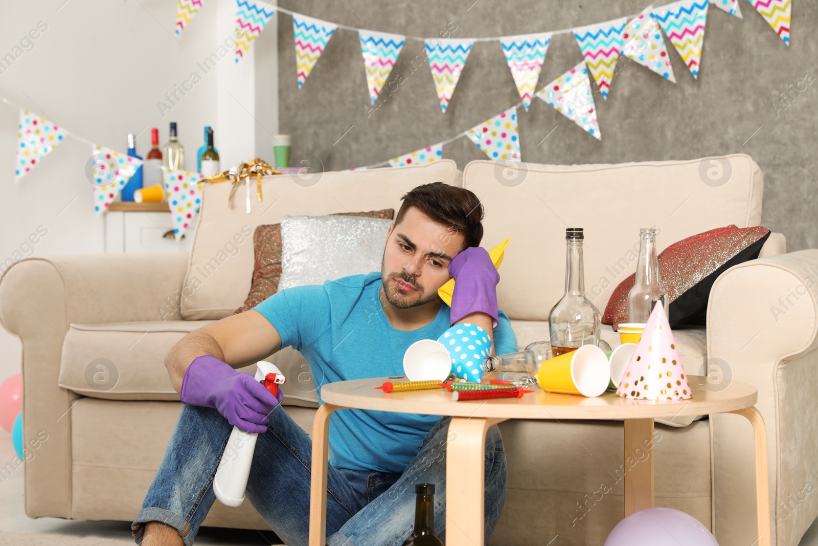 Photo of Tired young man with bottle of detergent and rag in messy room after party