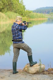Man aiming with hunting rifle near lake outdoors