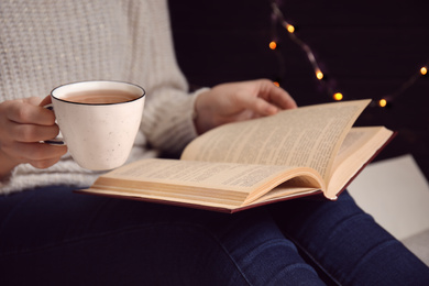 Woman with cup of coffee reading book at home, closeup