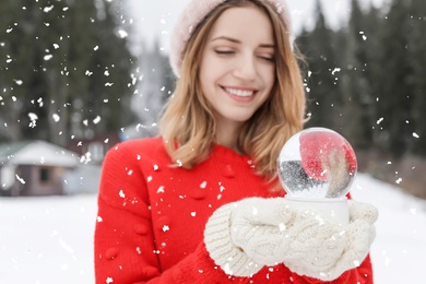 Young woman in knitted clothes holding snow globe outdoors. Winter vacation