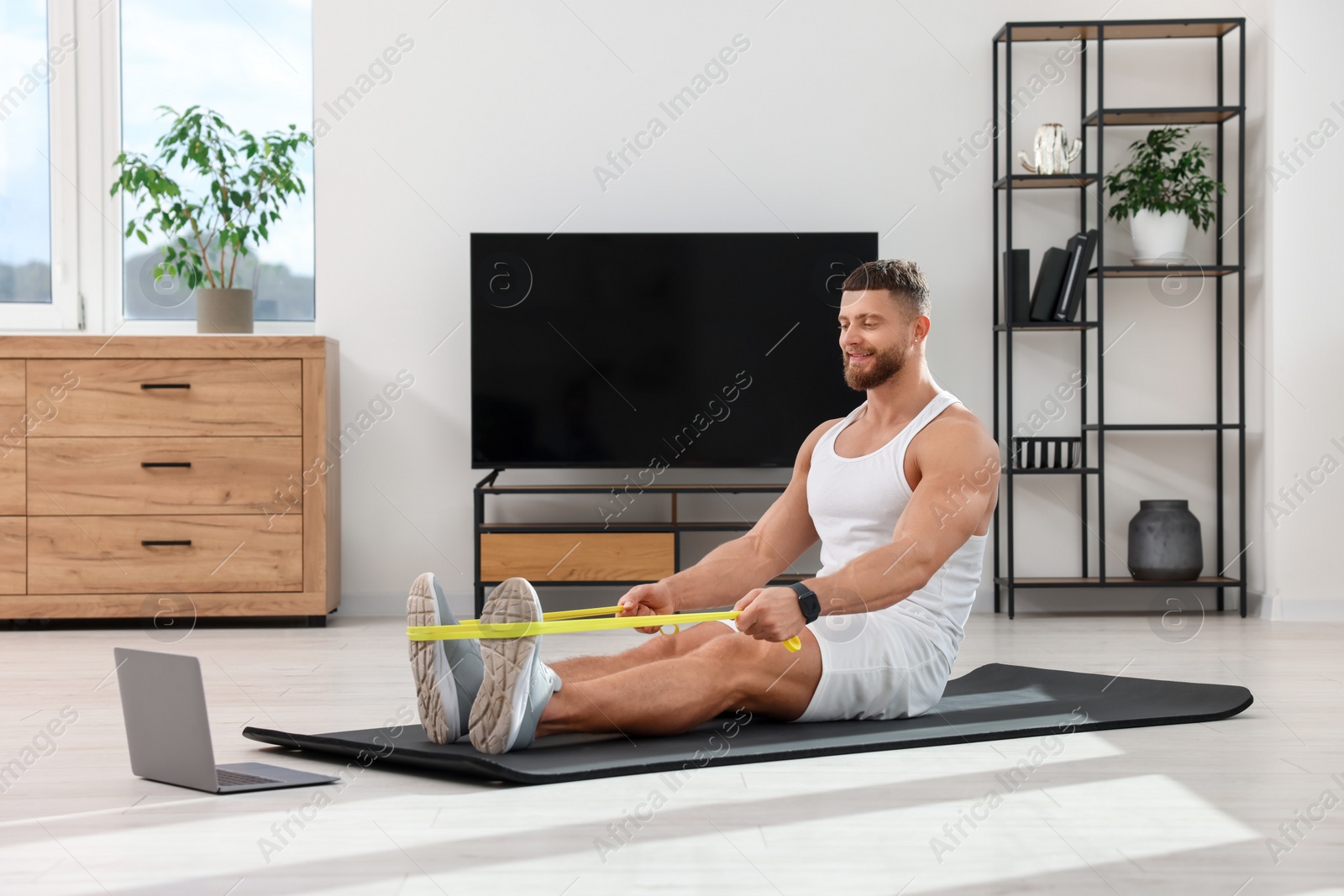 Photo of Muscular man doing exercise with elastic resistance band near laptop on mat at home