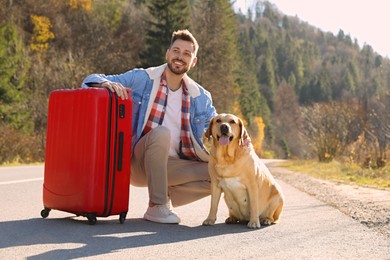 Happy man with adorable dog and red suitcase on road. Traveling with pet