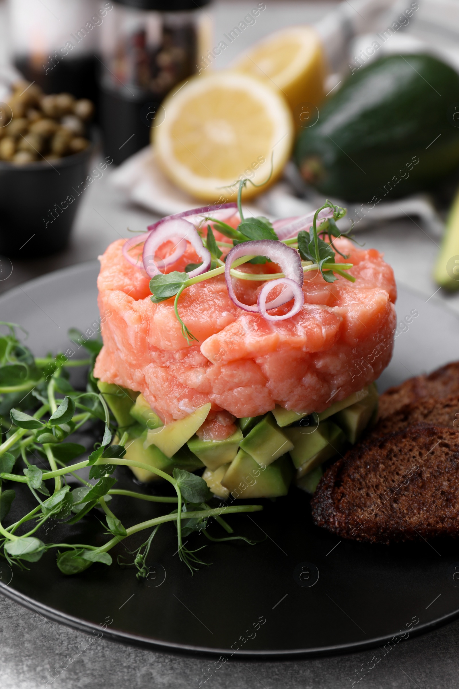 Photo of Tasty salmon tartare with avocado, microgreens and croutons on grey table, closeup