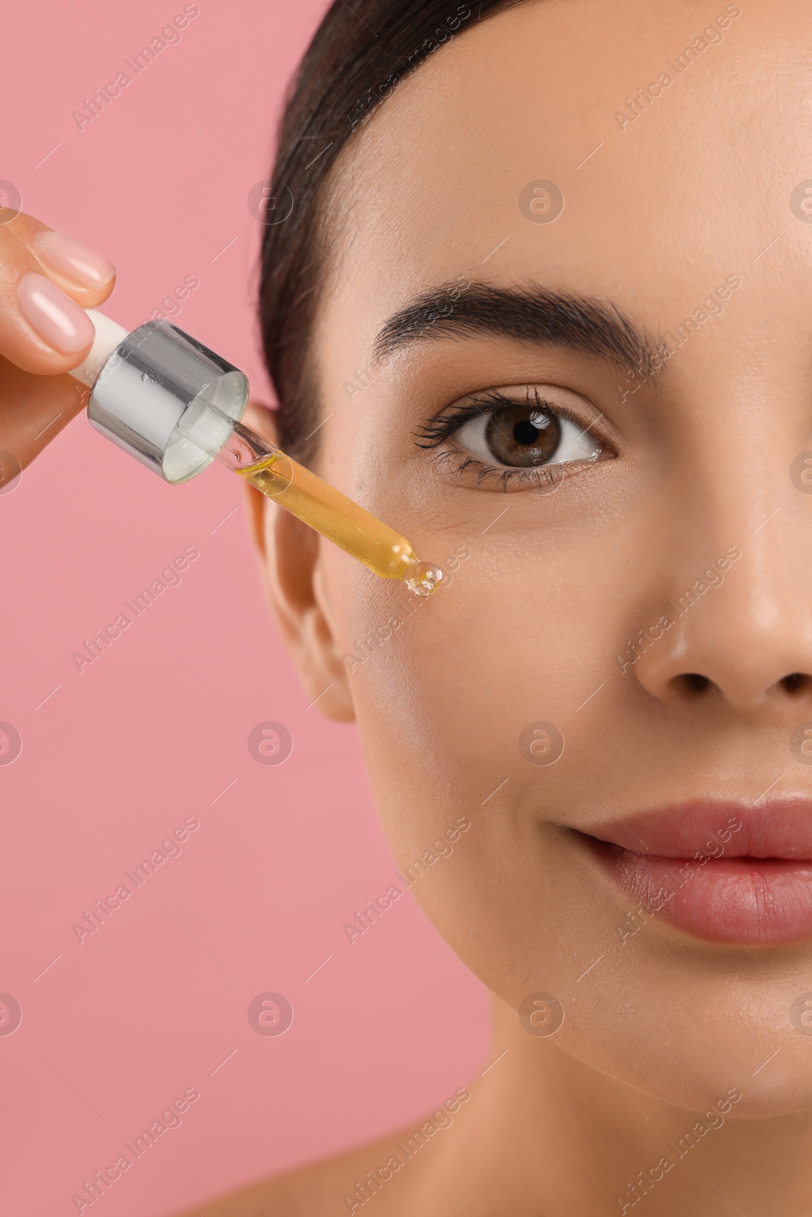 Photo of Beautiful young woman applying serum onto her face on pink background, closeup