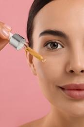 Beautiful young woman applying serum onto her face on pink background, closeup