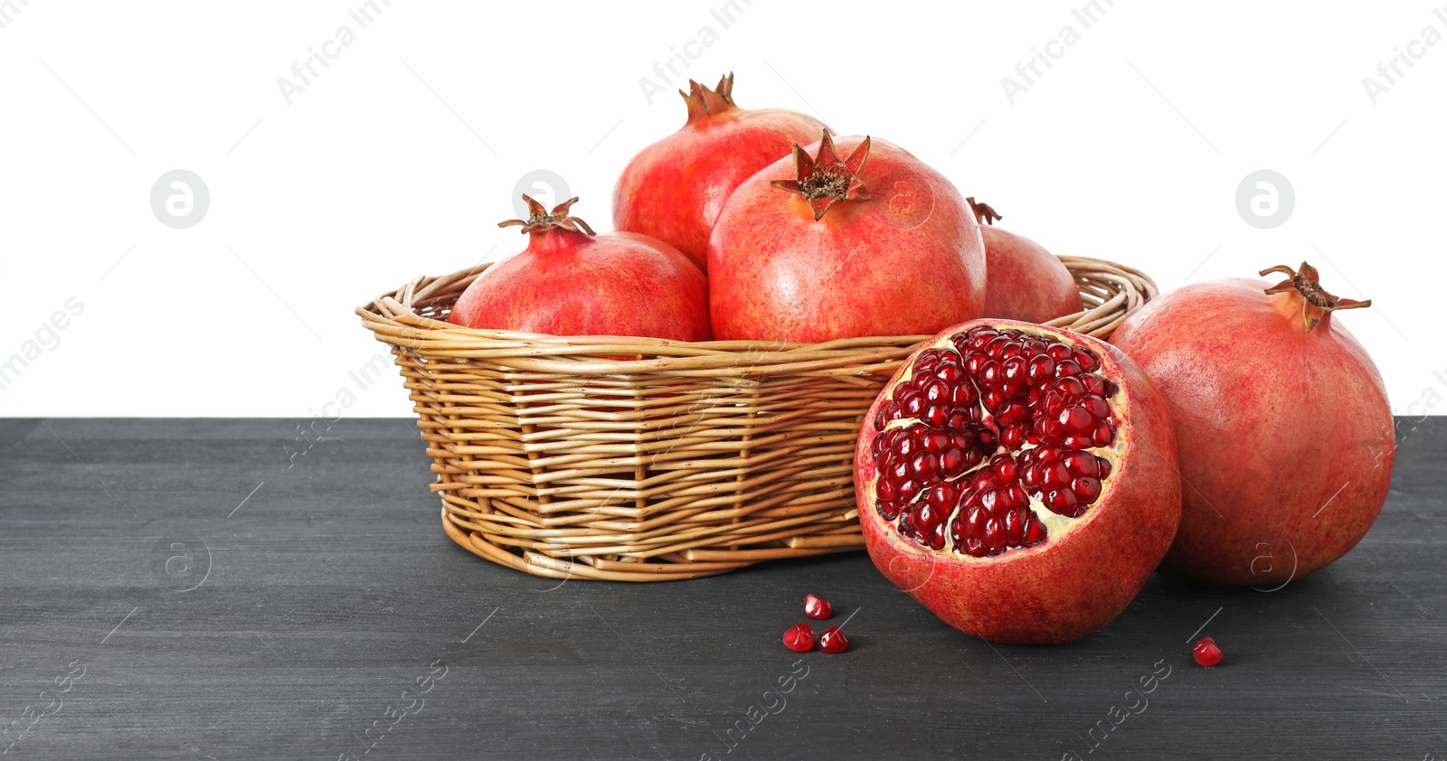 Photo of Fresh pomegranates in wicker basket on black wooden table against white background, space for text