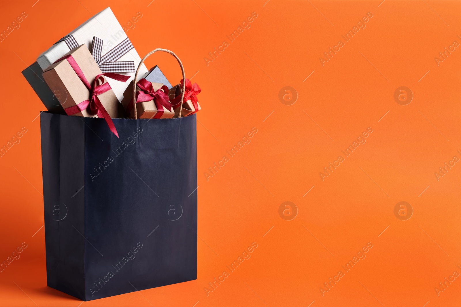 Photo of Dark blue paper shopping bag full of gift boxes on orange background. Space for text