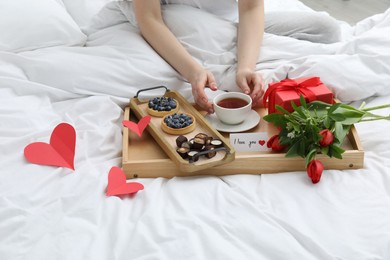 Photo of Tasty breakfast served in bed. Woman with tea, desserts, gift box, flowers and I Love You card at home, closeup