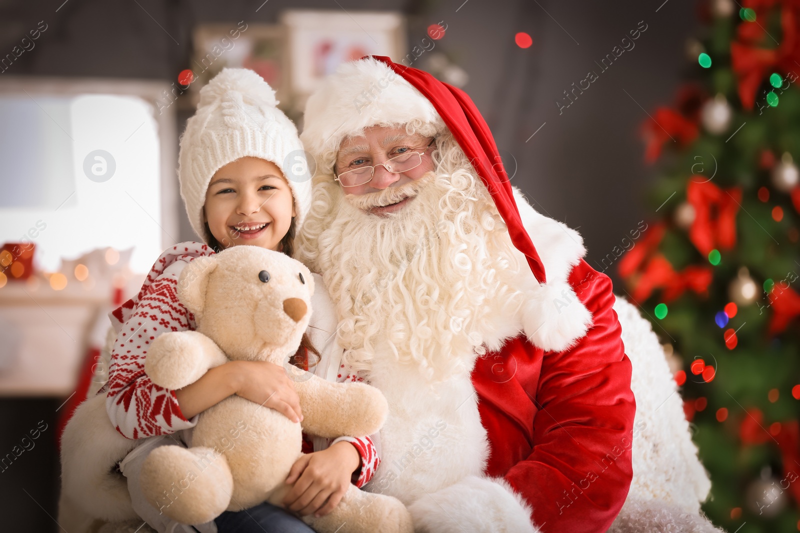 Photo of Little girl with teddy bear sitting on authentic Santa Claus' lap indoors