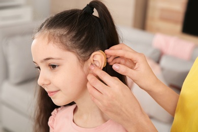 Photo of Young woman putting hearing aid in daughter's ear indoors