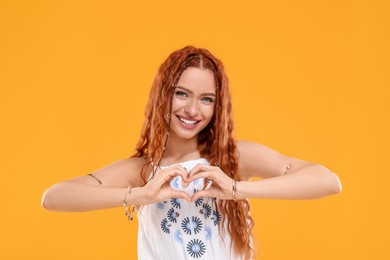 Beautiful young hippie woman making heart with hands on orange background