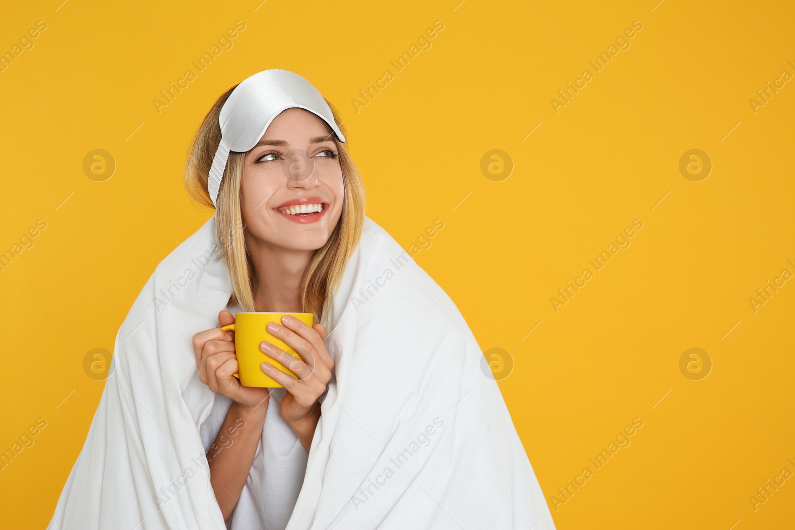 Photo of Young woman in sleeping mask wrapped with blanket holding cup on yellow background. Space for text