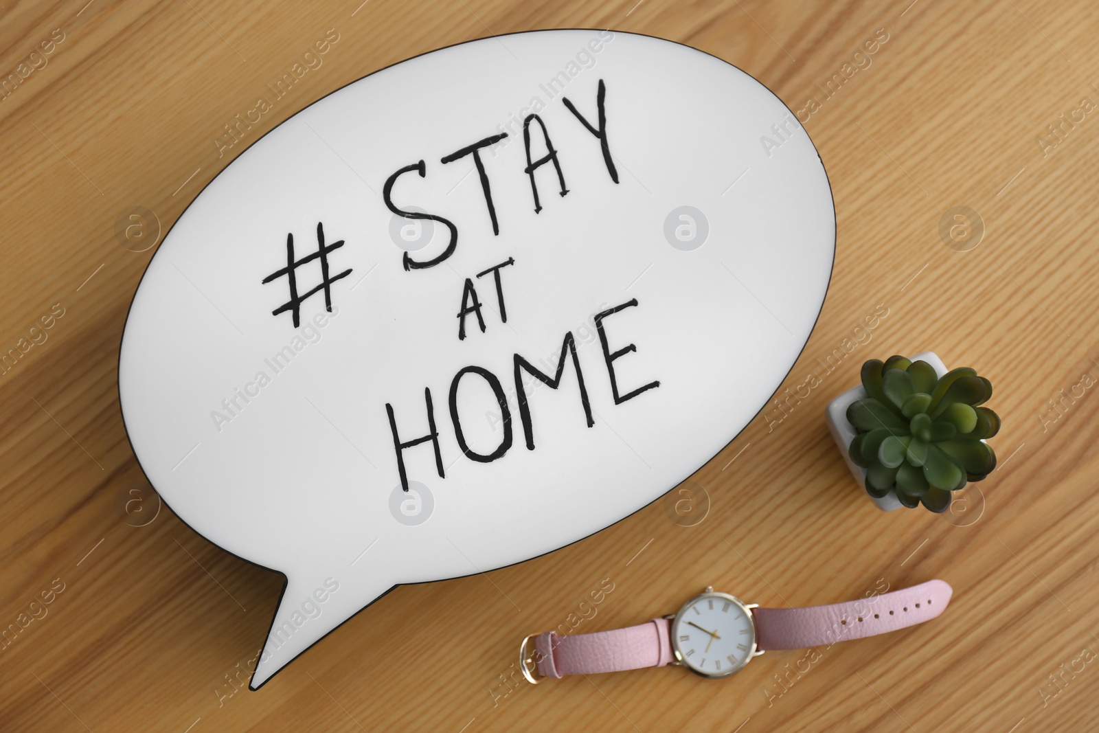 Photo of Houseplant, wristwatch and speech bubble with hashtag STAY AT HOME on wooden background, flat lay. Message to promote self-isolation during COVID‑19 pandemic