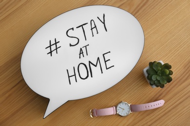 Photo of Houseplant, wristwatch and speech bubble with hashtag STAY AT HOME on wooden background, flat lay. Message to promote self-isolation during COVID‑19 pandemic