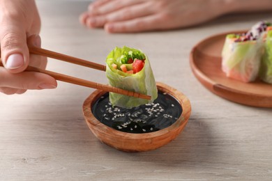Woman dipping delicious spring roll wrapped in rice paper into soy sauce at white wooden table, closeup