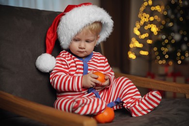 Photo of Cute little child in Santa hat with ripe tangerines in armchair at home