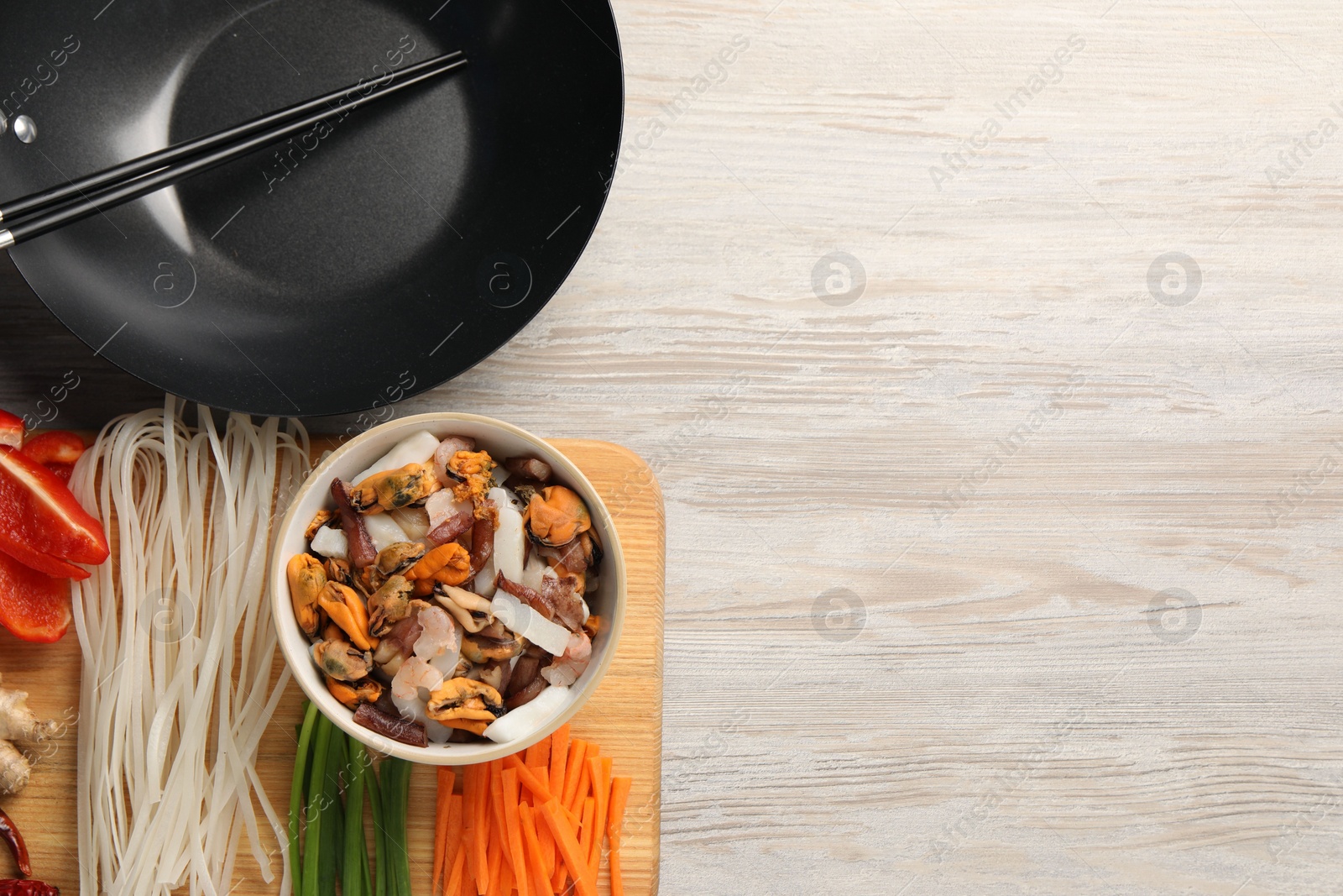 Photo of Black wok, chopsticks and board with products on light wooden table, top view. Space for text