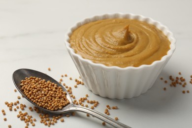 Photo of Tasty mustard sauce in bowl and spoon with dry seeds on white marble table, closeup