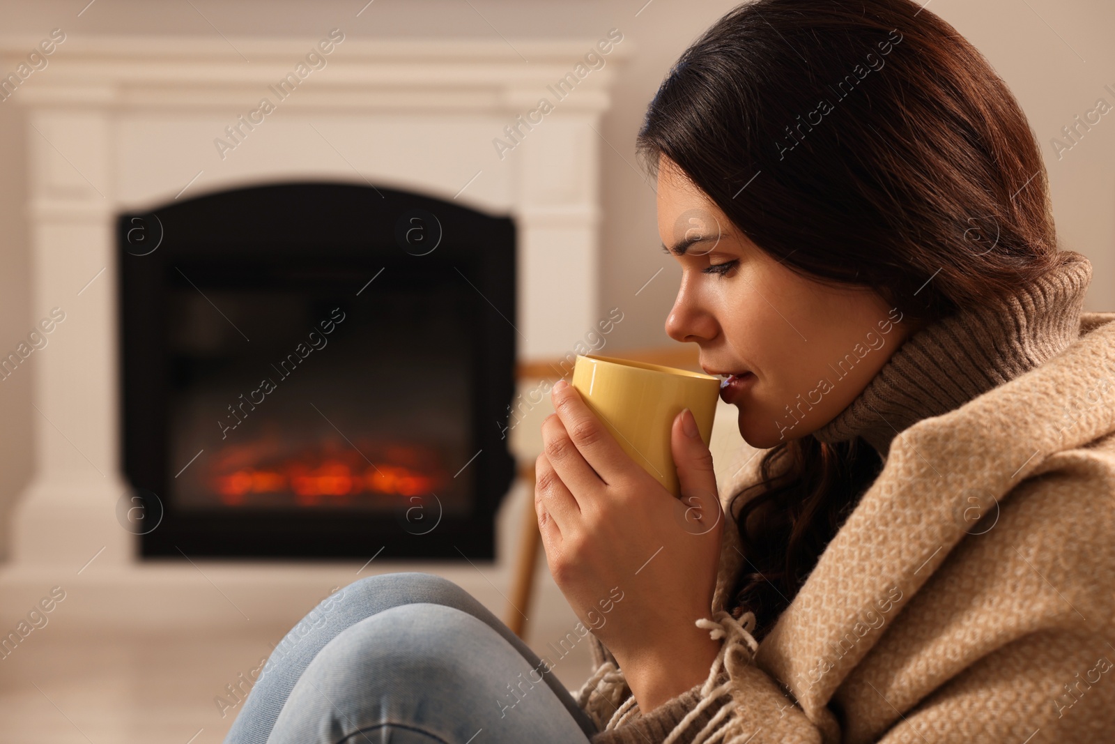 Photo of Young woman with cup of tea relaxing near fireplace at home. Space for text