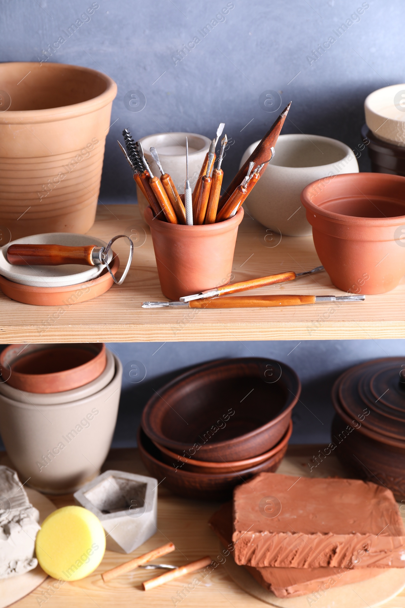 Photo of Set of different crafting tools and clay dishes on wooden rack in workshop, closeup