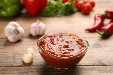 Delicious adjika sauce in glass bowl and ingredients on wooden table, closeup