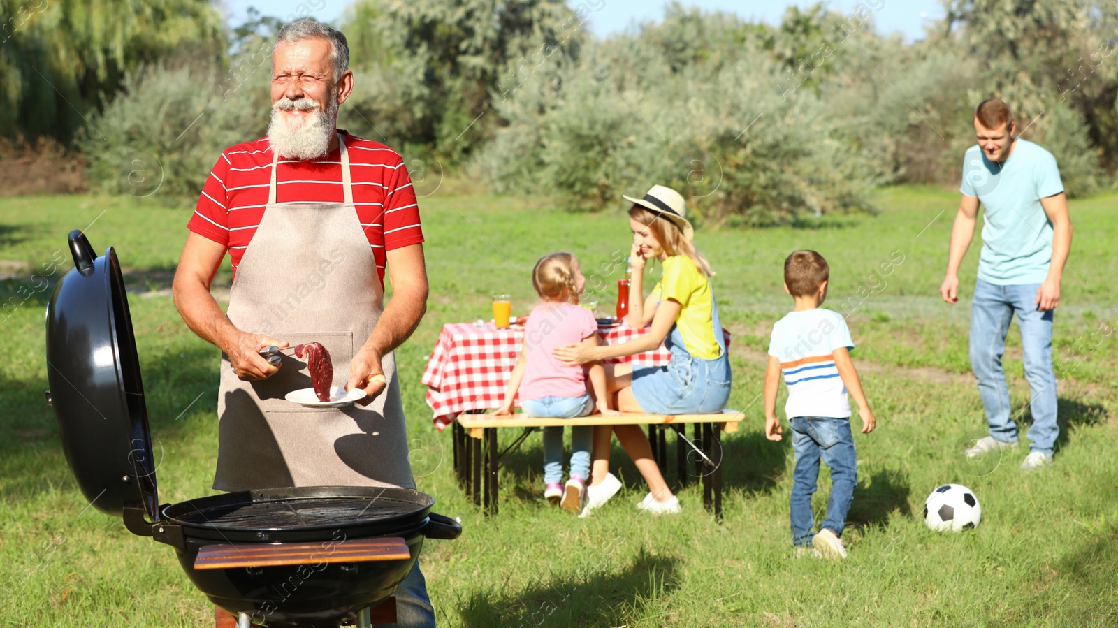 Photo of Happy senior man with meat at barbecue grill and his family having picnic in park