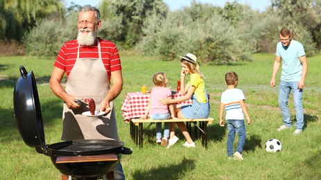 Photo of Happy senior man with meat at barbecue grill and his family having picnic in park