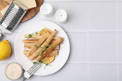 Plate with baked salsify roots, lemon, fork and sauce on white tiled table, flat lay. Space for text