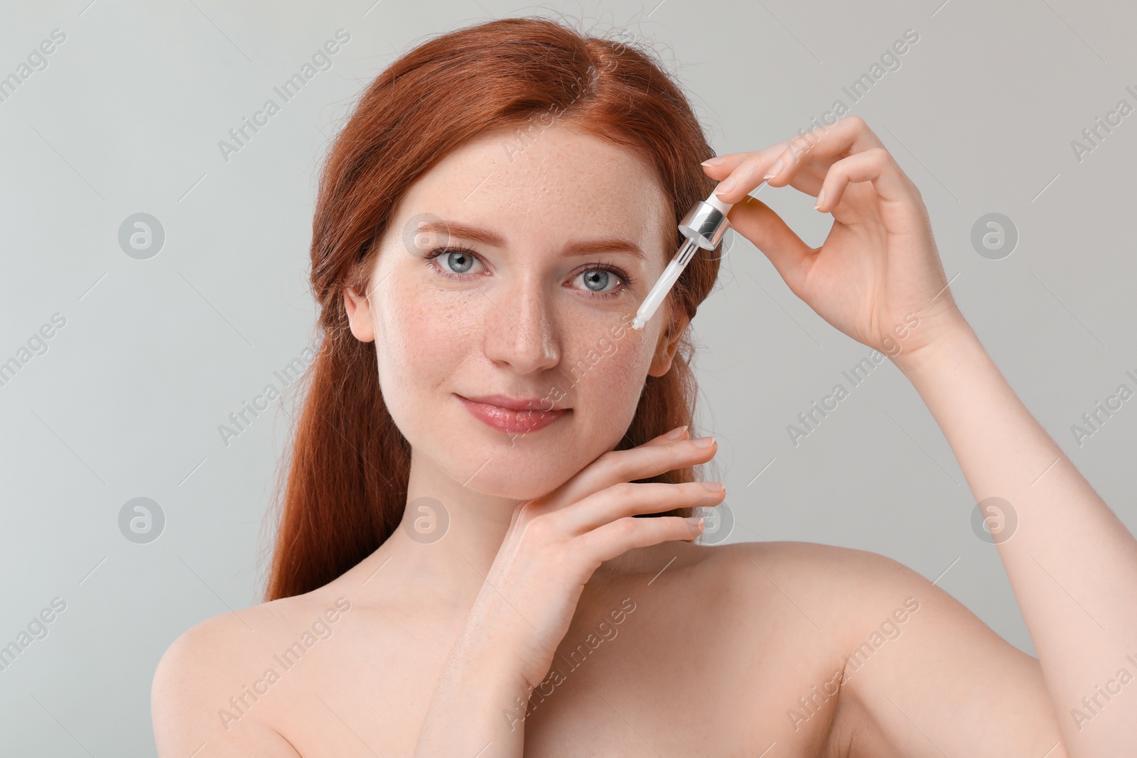 Photo of Beautiful woman with freckles applying cosmetic serum onto her face against grey background, closeup