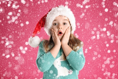 Image of Cute child in Santa hat under snowfall on pink background. Christmas celebration