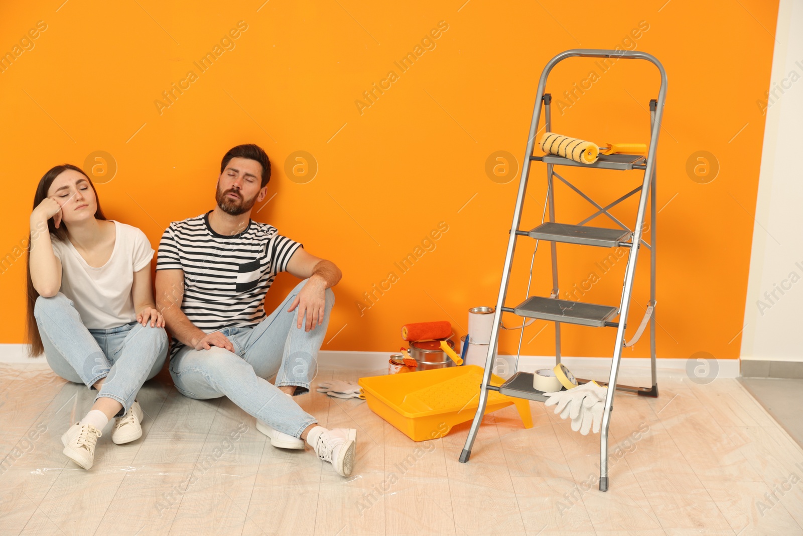 Photo of Tired designers sitting on floor and painting equipment near freshly painted orange wall