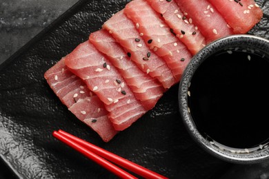 Photo of Tasty sashimi (pieces of fresh raw tuna with sesame seeds) and soy sauce on black plate, flat lay