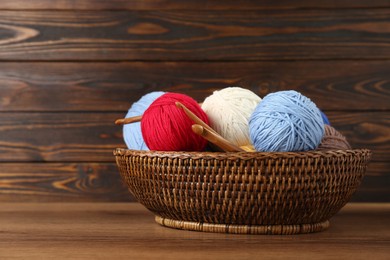 Photo of Wicker basket with clews of colorful knitting threads and crochet hooks on wooden table