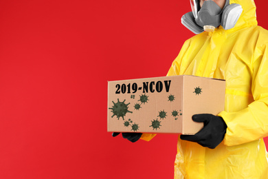 Man in chemical protective suit holding cardboard box on red background, closeup view with space for text. Coronavirus outbreak