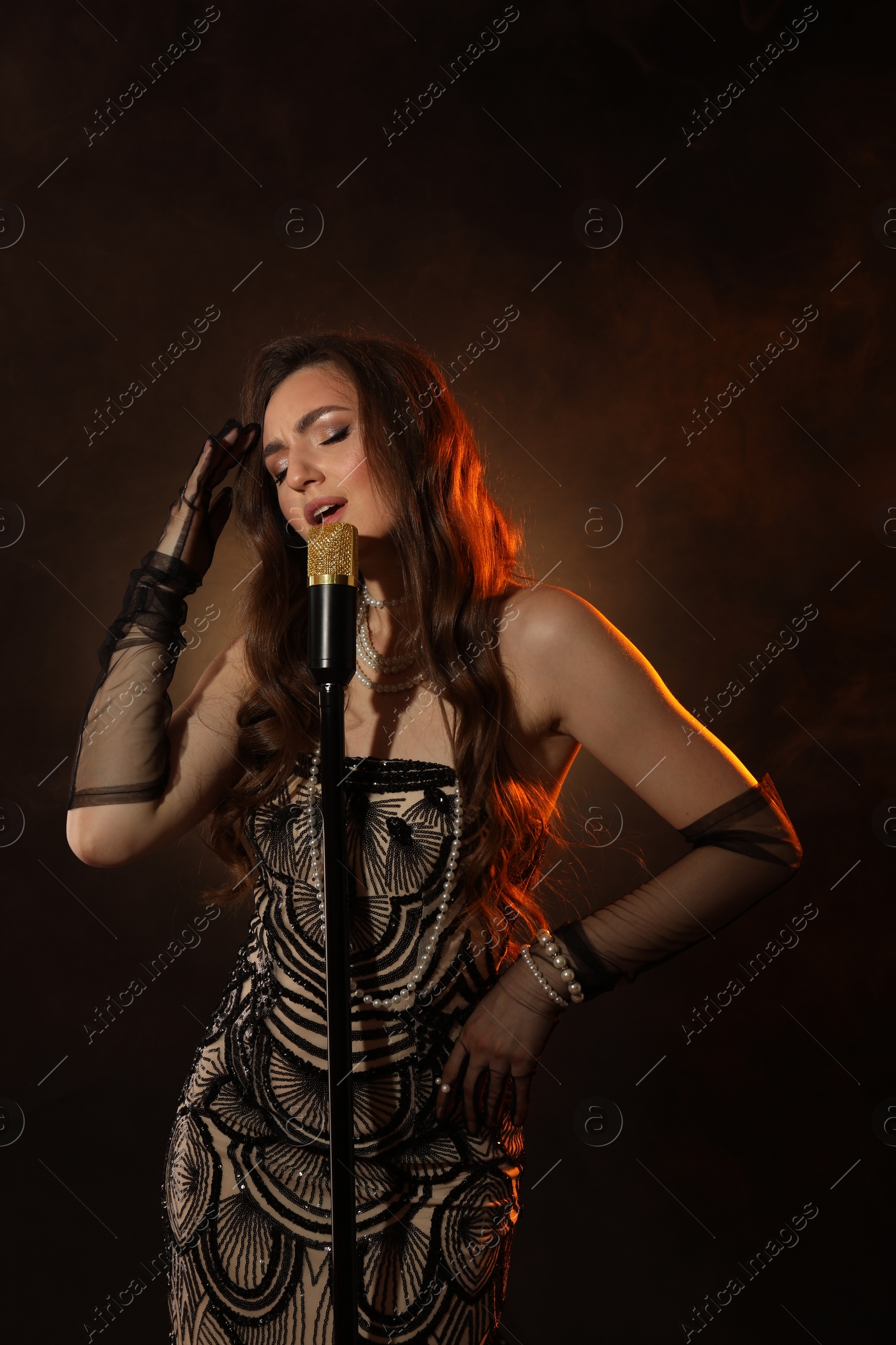 Photo of Beautiful young woman in stylish dress with microphone singing on dark background with smoke