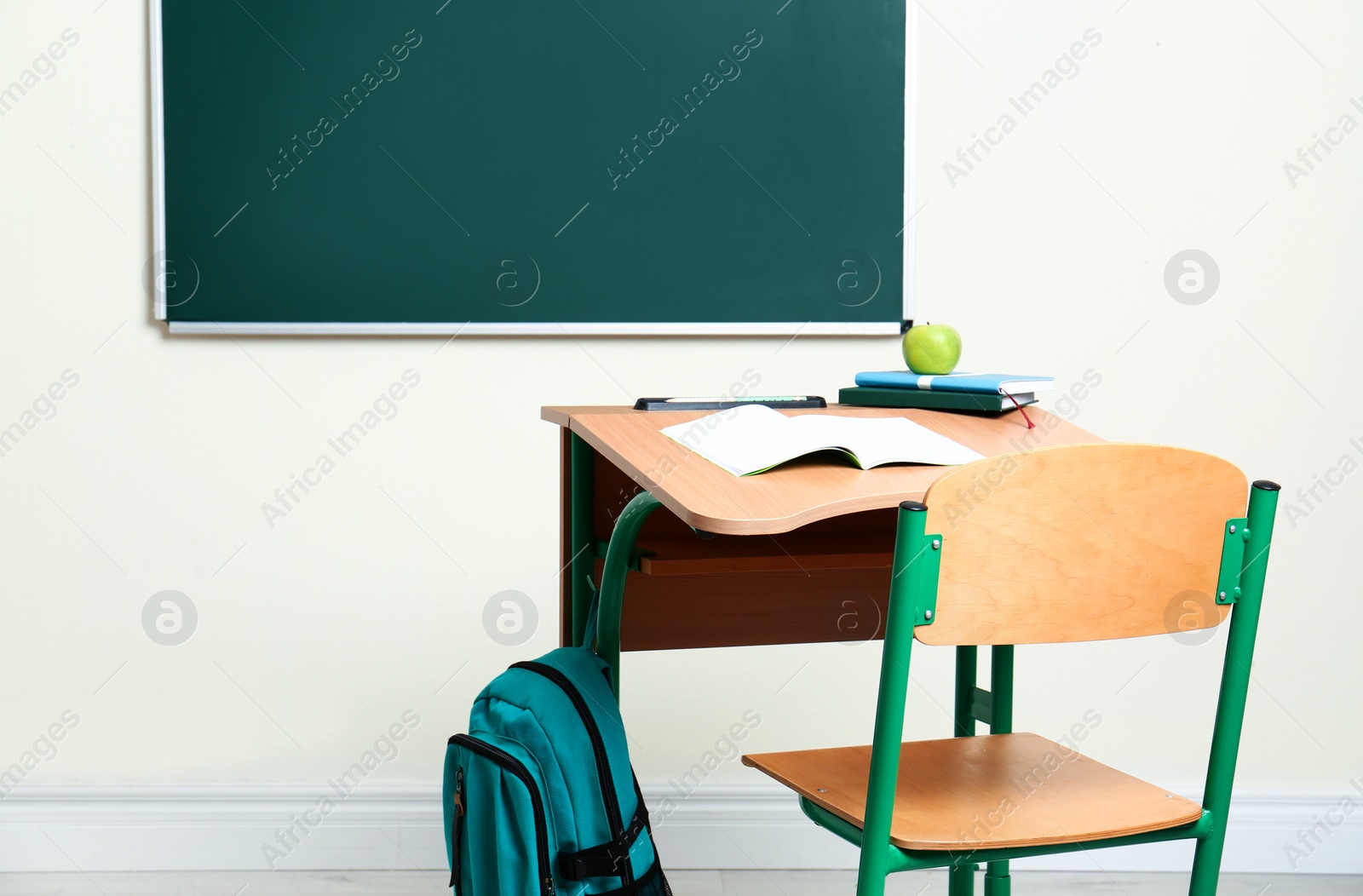 Photo of Wooden school desk with stationery, apple and backpack near chalkboard in classroom