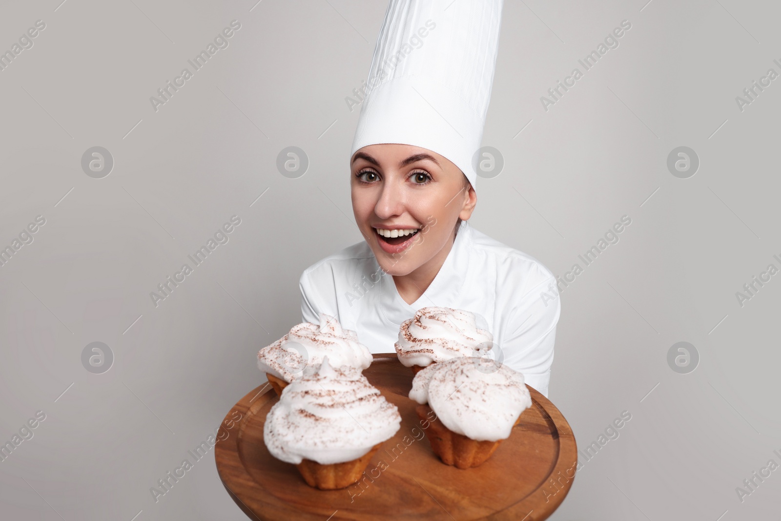 Photo of Happy professional confectioner in uniform holding delicious cupcakes on light grey background