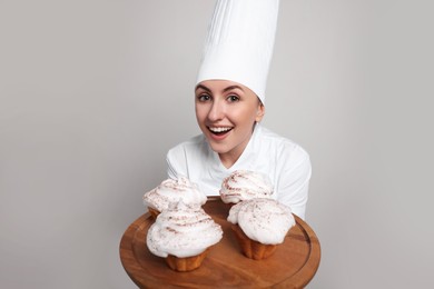 Happy professional confectioner in uniform holding delicious cupcakes on light grey background
