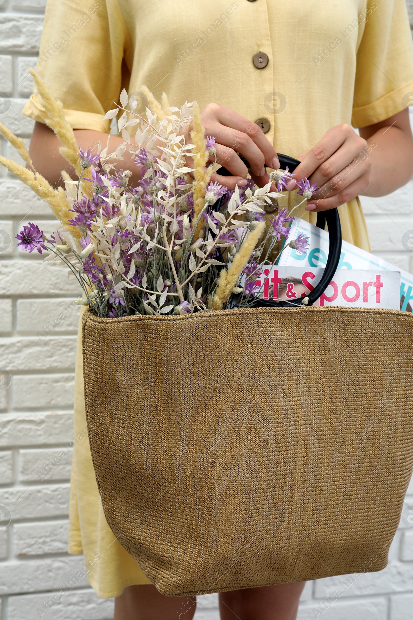 Photo of Woman holding beach bag with beautiful bouquet of wildflowers and magazines near white brick wall, closeup