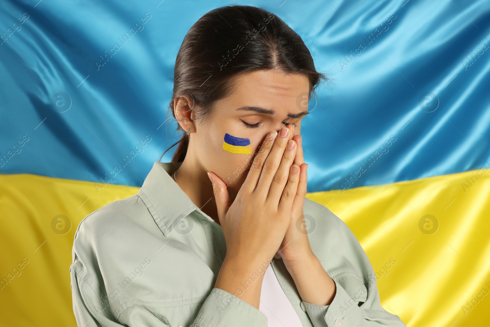 Photo of Sad young woman with clasped hands near Ukrainian flag